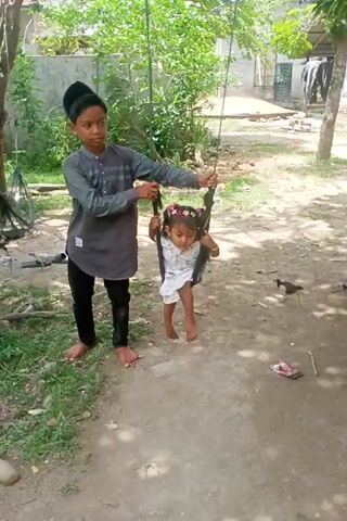 A young boy, dressed in a grey shirt, black pants, and a black cap, is manually swinging a younger girl on a makeshift swing made from rope and a seat. The girl, wearing a white dress and a flowered headband, lies in the swing, barefoot. Both are outside, on a dirt ground partially covered in grass, under a tree. There is a building and some greenery in the background. The scene appears to be a candid shot of the two children playing.