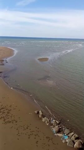 The video shows an aerial view of a beach and the sea. The water is a murky green-brown color and appears to be shallow near the shore. There are several sandbars visible in the water. On the beach, there is a pile of rocks and some blue plastic debris. The sky is blue with a few white clouds. The sound of a child crying can be heard.