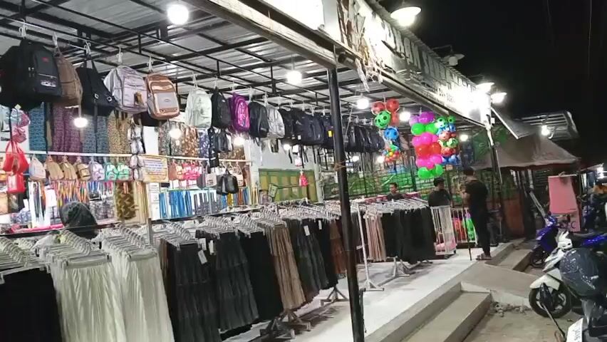 The video shows an outdoor market stall at night. The stall is filled with various items for sale, including backpacks, clothing such as skirts and t-shirts, and bags. There are also some colorful balloons hanging in the background. Several people are visible in the video, including customers browsing the merchandise and possibly vendors. The market appears to be well-lit with artificial lights. Scooters are parked near the stall. The overall impression is of a busy, vibrant night market.