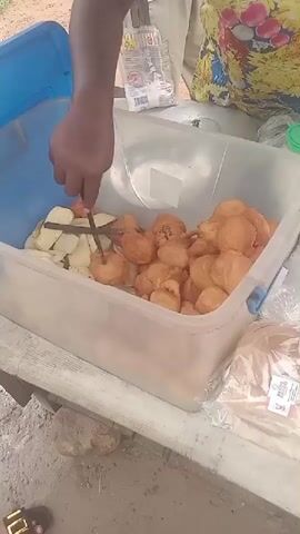The video shows a vendor preparing food from a large container at an outdoor stand. The container holds fried balls and sliced plantains. The vendor uses a wooden stick to poke the food. The vendor also cuts open a plastic bag containing sliced bread. Other items on the table include a jar with a green lid, a pack of instant noodles, and more bread in plastic bags. The vendor's hands are visible as they manipulate the food items. The vendor wears a patterned yellow and blue top. A person wearing sandals is visible at the bottom of the frame. The setting appears to be a sandy or dusty outdoor area.