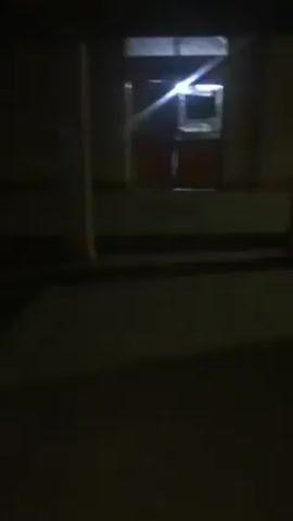 The video depicts a dimly lit hallway with beige walls and a gray, tiled floor.  On one side, there are several white doors with silver handles, interspersed with silver vertical cabinets that appear to be refrigerators or storage units.  On the opposite side, the wall is mostly flat and beige, with a single red fire extinguisher mounted.  At the far end of the hallway, light streams in from an open doorway, revealing a glimpse of bright colors and shapes. The camera moves forward, progressing down the hallway. No people are visible in the immediate foreground of the hallway, but what appears to be one person can be seen in the distance.