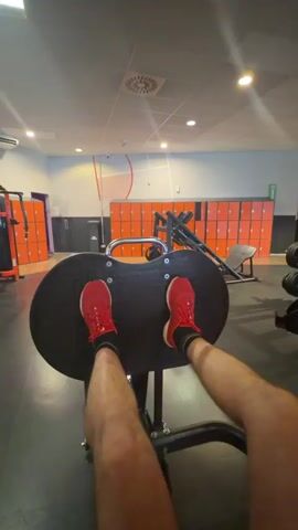 The video shows a first-person perspective of a person's legs, wearing red sneakers and black socks, performing a leg press exercise in a gym. The person is sitting on a black leg press machine with their feet placed on a large black platform. Their knees are bent, and they are extending their legs upwards to push the platform away. The gym environment is visible in the background, with orange lockers lining the wall, gym equipment, and fluorescent lighting. The video captures multiple repetitions of the leg press motion.