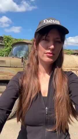 A woman wearing a black baseball cap and a black zip-up jacket is standing in front of a rusty, olive-green truck on a sunny day. The truck appears to be an older model, possibly a Soviet-era vehicle, with a water tank attached to the back. The woman has long, brown hair and is looking directly at the camera. The background shows a slightly overgrown area with some stone structures and trees. The scene suggests an outdoor, rural or semi-rural setting.