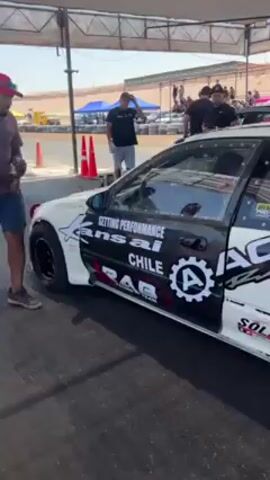 Here's a detailed description of the video:

The video shows a white Honda Civic race car, heavily modified with decals and racing parts, at a drag strip. The car has a roll cage visible through the window, indicating it's designed for racing. Decals such as "ANSIA Performance Chile," "CHULZ Renewable," and others are visible on the car's body. A large exhaust outlet is present at the rear. The car has a Japanese license plate.

Around the car, people are standing and observing. Some are wearing t-shirts and caps, suggesting they are part of the pit crew or spectators. The setting is an outdoor race track on a sunny day, with tents and support vehicles visible in the background.