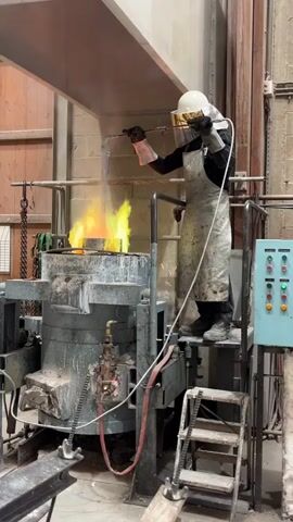 The video shows a person in protective gear working with a large industrial furnace. The person is wearing a white hard hat with a gold visor, a long light-colored apron, black gloves, and black clothing. They are standing on a metal platform next to the furnace. The furnace is large and cylindrical, with metal structures and pipes surrounding it. Flames are visible inside the furnace, indicating it is in operation. The person is holding a long metal rod and appears to be manipulating something inside the furnace. The environment looks like a metalworking or manufacturing facility, with a brick wall visible in the background and various industrial equipment nearby.