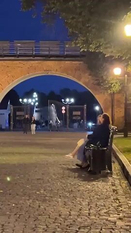 The video captures an evening scene at a park. The ground is paved with cobblestones, and a stone archway looms in the background. Two people are seated on a bench in the foreground, facing away from the camera. One person is wearing a dark jacket and the other a blue dress. Beyond the archway, a set of ornate gates is visible, illuminated by streetlights. Several figures are walking near the gates. A street lamp is present on the right side of the frame. Trees with green foliage are seen at the top of the image, and the sky is a deep blue, indicating dusk or nighttime.