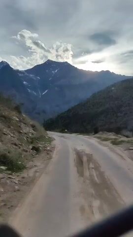 A POV shot from a vehicle traveling on a paved road winding through a mountainous landscape. The road is in fair condition with some potholes. To the left of the road is a rocky incline with sparse vegetation. To the right, the terrain slopes down into a valley covered with trees. In the background, majestic mountains with snow-capped peaks dominate the skyline. The sky is partly cloudy, with sunlight breaking through the clouds and casting rays onto the mountains. The overall atmosphere is serene and depicts a journey through a scenic natural environment.