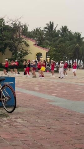 A group of about a dozen women are participating in an outdoor exercise class in a plaza. They're dancing in synchronized motion, some wearing red outfits with hats, while others sport a mix of colors. Behind them, there's a building painted yellow and pink, surrounded by palm trees and other greenery. The plaza is paved with red bricks, and in the foreground, a bicycle leans against a blue object.