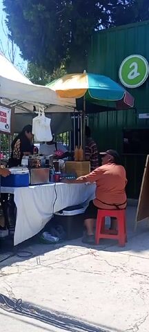 Here is a description of the video:
The video shows a small outdoor food stand. There are four people around the stand. One is sitting on a red stool facing the stand. There is a white tent over the stand. Next to the stand is a green building with a large green "a" logo on it. There is a small umbrella near the green building. A man in a purple shirt is placing a sign next to the stand.