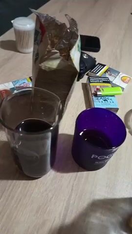 The media file shows a close-up of a wooden table with various items on it. In the foreground, there are two glasses. The glass on the left contains a dark liquid, possibly wine or a soft drink. The glass on the right is purple and also contains a dark liquid. A bottle of clear liquid, likely spirits, is being poured into the purple glass. Behind the glasses, there's an open bag of chips or snacks, a pack of cigarettes, and a wallet. A white container with toothpicks is also visible in the background. The lighting suggests it could be daytime or evening indoors. The overall scene depicts a casual gathering or a moment of relaxation.

Visible text includes:
- "CAMEL" and "MENTHOL" on a cigarette pack.
- "POGUES" and "IRISH WHISKEY" on the purple glass.
- Text on the chip bag is partially obscured but appears to be in a foreign language, possibly Bulgarian given the location context.
- A price tag or label is visible on a small object behind the chip bag, but the text is illegible.