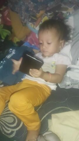 A child is lying on a bed, looking at a smartphone. The child is wearing a white t-shirt and yellow pants. The bed appears to be covered in blankets and clothing. The lighting in the room is dim.