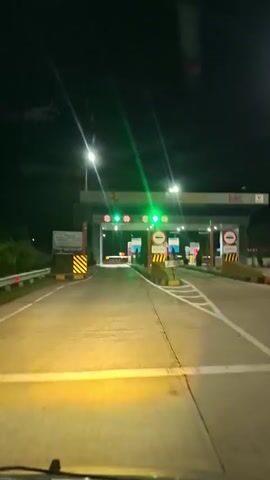 The video, captured from a car's perspective, shows a vehicle approaching a toll booth at night. The road ahead is illuminated by the car's headlights, casting a yellow glow. Overhead, bright streetlights cast a white light on the scene. The toll booth structure is a large, grey building with several lanes. Green arrows are visible above two of the lanes, indicating they are open, while other lanes show red crosses, indicating they are closed. A 'no entry' sign is visible to the right of the open lanes. On either side of the road leading to the toll booth, there are yellow and black striped barriers. To the left, a sign is visible, possibly displaying information for drivers. The overall scene suggests a nighttime drive on a highway leading to a toll plaza.