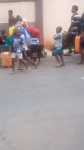 The video is filmed in a portrait orientation and is noticeably shaky and blurry, indicating it was likely recorded handheld with a low-resolution camera. The setting appears to be an outdoor, possibly urban or semi-urban environment, with a light gray paved surface (likely asphalt or concrete) serving as the ground.

At the beginning of the video (0:00-0:01), a group of about 5-7 individuals are visible in the mid-ground, standing and milling about. They are casually dressed in t-shirts and shorts of various colors, including red, blue, and light tones. Hints of green foliage can be seen on the left side of the frame, suggesting trees or bushes in the background.

From 0:02 to 0:05, a man walks into the foreground and becomes the central focus. He is wearing a white short-sleeved t-shirt with a large graphic on the front featuring the number "51" in prominent red lettering, possibly accompanied by other blue text that is illegible due to the blurriness. He also wears dark-colored shorts and light green flip-flops. He has dark hair and skin. As he walks, he looks towards the camera and raises his right hand, making a gesture that appears to be a peace sign (V-sign) or pointing with two fingers. A light green wristband is visible on his right wrist. He maintains eye contact with the camera as he passes by.

In the background throughout this segment, other blurry figures of people can be seen, some standing and others possibly sitting, contributing to the impression of a casual gathering or a public space. The lighting suggests it is daytime, with no strong shadows indicating early morning or late evening. No specific text on signs or buildings is discernible in the video.