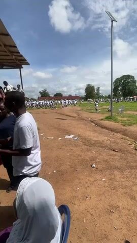 The video shows a group of young people, many wearing white shirts, white shorts, and green hats, standing in an outdoor area with a dirt ground. Some are walking, while others are standing in formation or small groups. There are buildings with red roofs in the background, and trees and grass are visible. Several plastic water bottles are scattered on the ground. A solar-powered lamp post is also present. The overall impression is of an organized outdoor event or training.