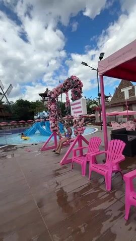 This is a vertical video shot in what appears to be a water park. In the foreground, there is a pink swing set adorned with flowers, and a person is sitting on it. To the right of the swing, there are two pink chairs. Beyond the swing is a swimming pool with blue water, and further back are pink umbrellas and lounge chairs. In the distance, there are water slides and structures resembling windmills. The sky is cloudy with patches of blue. The video captures a person swinging, the pool area, and the surrounding water park attractions.