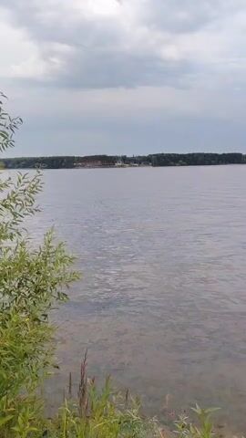 This video captures a serene view of a lake on a cloudy day. The foreground features lush green grass and wildflowers, suggesting a natural, outdoor setting. The lake stretches out to the horizon, where a dense line of trees forms a dark, continuous backdrop. In the distance, across the water, some structures are visible, possibly part of a resort or development.

The activity taking place is subtle. Several buoys, marked in bright orange, are scattered across the water, indicating a designated area for swimming or water activities. Further out, a couple of people can be seen on what appears to be a small boat or raft, enjoying their time on the lake. The overall mood is peaceful and tranquil, amplified by the calm waters and overcast sky. The time of day is difficult to pinpoint precisely due to the clouds, but it appears to be daytime. The presence of the buoys and distant structures hints at a recreational area, consistent with the provided location context of "Туристический Пансионат «Клязьминское водохранилище», Russia." There is no visible text in the video.