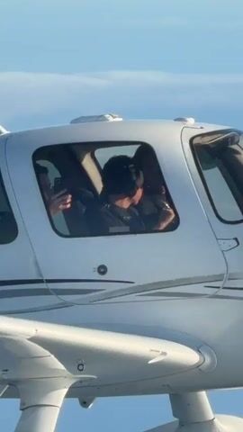 A close-up, exterior shot captures a white airplane in mid-flight against a bright blue sky. The image focuses on the cockpit windows, revealing two individuals inside. The person in the left seat is wearing headphones and appears to be holding a mobile phone, possibly taking a picture or recording. The person in the right seat, who is likely the pilot, is also wearing headphones and looking forward, seemingly engaged in flying the aircraft. The plane has a sleek design with grey pinstripes along the fuselage and two prominent white landing gear pods visible beneath the wings. The overall impression is one of peaceful flight and shared experience.