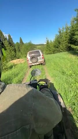 From the perspective of an ATV rider, the video shows a grassy field with a white SUV stuck in the distance. The ATV is moving along a narrow dirt path through the tall green grass. There are several young evergreen trees scattered throughout the field, and the sky is a bright blue with a few white clouds. The ATV rider is wearing a green jacket, yellow gloves, and a neon green and black helmet. A cell phone is mounted on the handlebar of the ATV.