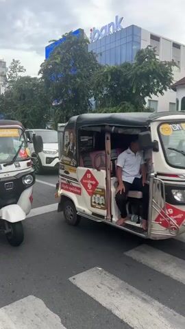 The video shows a street scene with several vehicles, including tuk-tuks and cars. In the foreground, there are two tuk-tuks facing forward. The one on the left is white with red accents and has a person in the driver's seat. The one on the right is also white with red accents and appears to have at least two people inside. Both tuk-tuks have advertisements and logos on them. In the background, a white car is visible in traffic, and behind it, a modern building with the word "abank" in blue lettering is prominently displayed. Trees line the street in front of the building. The sky is overcast. The camera is positioned at street level, capturing the vehicles and the urban environment. The video appears to be shot from a moving vehicle or while standing still in traffic.