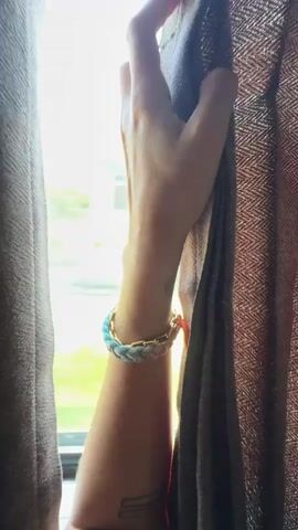 The video shows a person's arm and hand holding onto a curtain inside a moving vehicle. The hand is wearing a braided blue and white bracelet with a gold clasp and a small orange charm. Through the window, blurred scenery of trees and hills can be seen, indicating the vehicle is in motion. The sky is blue with some white clouds. The scene appears to be daytime.
