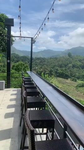 The video shows a view from an outdoor balcony or patio. In the foreground, there is a long, dark counter with several woven bar stools lined up along it. A string of exposed light bulbs hangs overhead, stretched across the frame. Beyond the counter, a lush green landscape unfolds with rolling hills covered in dense forest. In the mid-ground, there are terraced fields, possibly rice paddies, and a few buildings. The sky is partly cloudy with a bright, diffused light, suggesting it is daytime. The overall impression is one of a scenic overlook, likely at a cafe, restaurant, or lodging with a view. There are no people visible in the video.