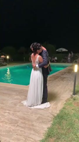 A bride and groom are captured in a close embrace by the side of a glowing turquoise swimming pool at night. The bride, in a flowing white wedding gown with an open back, is facing away from the camera, her head tilted back as she kisses the groom. The groom, dressed in a dark suit, has his arms wrapped around her. The poolside is paved with light-colored stones, and the surrounding area is dimly lit by outdoor lamps and the pool's illumination. In the background, trees and foliage are visible, silhouetted against the dark night sky. A few figures are seated in the background near a white umbrella, and a light fixture is prominent on the right side of the frame. The overall atmosphere is intimate and romantic.