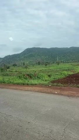 The video shows a view from a moving vehicle looking out at a rural landscape. The foreground features a cracked asphalt road with some green vegetation on the side. Beyond the road, a large, verdant field stretches out, dotted with scattered trees. In the background, a majestic green mountain range rises under a cloudy sky. The overall scene depicts a serene and natural environment.