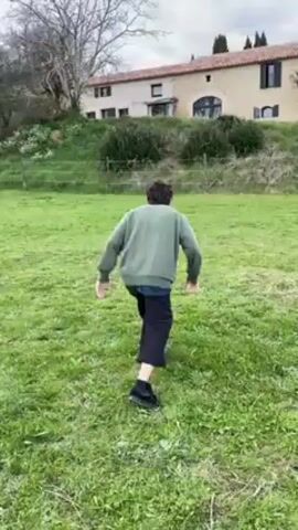 A person with dark, curly hair, wearing a green sweatshirt and dark pants and shoes, runs toward the camera across a grassy field, attempts a forward roll, and lands awkwardly in a seated position. They sit cross-legged, facing away from the camera, in the center of the frame. A light-colored stone building with dark shutters is visible in the background, situated on a slight hill. A bare tree stands to the left of the building. A wire fence runs along the middle ground of the scene. The sky is overcast.
