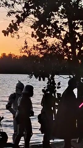 A group of people in Star Wars costumes are gathered on a sandy shoreline near a body of water. The sun is setting, casting a warm orange glow across the sky and reflecting on the water. Among the costumed individuals are at least two stormtroopers, one of whom appears to be a clone trooper. One person is dressed as Darth Vader, holding a pink lightsaber. Another individual is holding a similar pink lightsaber. The group is standing in a loose formation, facing the water or each other. The environment is natural, with trees and foliage visible in the foreground and background. A bicycle is partially visible in the lower left corner of the frame.