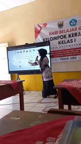 A woman wearing a black hijab and a patterned top is standing in front of a large interactive screen, holding a microphone. She is pointing at the screen, which displays numbers and images, including some animals. The screen appears to be part of an educational presentation, possibly for children, as there are images of ducks, frogs, and other animals alongside numbers and simple math equations. A banner with text in Indonesian hangs on the wall behind her, indicating a "LEARNING DAY" event for "CLASS 1." The room appears to be a classroom, with wooden tables covered in red cloths visible in the foreground. The lighting is warm, suggesting interior lighting.