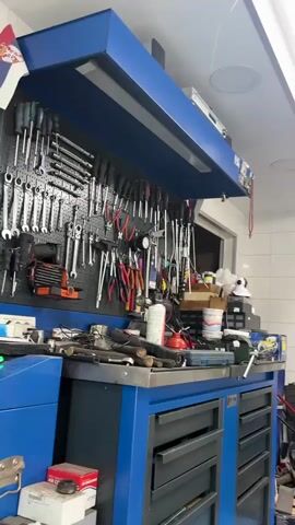 The video shows a car repair shop, beginning with a shot of a blue workbench cluttered with tools and equipment. The camera then pans right to reveal a blue car elevated on a yellow scissor lift. The front wheels are removed, exposing the brake rotors. The environment is a well-lit, tiled garage. The lift sits above a sunken service pit, visible through its grate. The car's front license plate is partially visible. A variety of equipment and supplies are scattered around the garage, including boxes, fluid containers, and a small refrigerator. Another car is visible outside the open garage door.