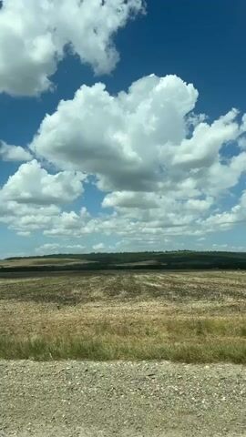 The video shows a rural landscape under a bright blue sky with fluffy white clouds. In the foreground, there is a gravelly roadside with some dry grass. The middle ground is a harvested field, appearing as stubble and soil. In the distance, a line of trees forms a dark horizon. A single traffic sign, a red triangle with a white border and a black symbol, is visible on the left side of the road. Towards the end of the video, a red tractor with attached farming equipment is seen working in the field, plowing or tilling the land. The overall scene depicts agricultural activity in a rural setting during the daytime.