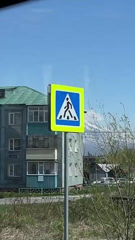 A video taken from a vehicle shows a snow-capped mountain range with clouds obscuring parts of the peaks. In the foreground, a light blue apartment building with balconies is visible on the left. Further into the background, there are several small, low-rise buildings, some appearing to be sheds or garages, and a line of parked cars. A traffic sign indicating "no overtaking" is prominently displayed on a pole in the mid-ground. The sky is clear and blue, suggesting daytime. The overall impression is of a rural or semi-urban setting with a striking natural landscape as a backdrop.