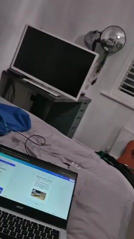 Here is a detailed description of the video:

The video presents a first-person perspective of a bedroom interior. The camera is handheld, resulting in some shakiness during the movement.

In the foreground, a laptop sits on a bed covered with a grey duvet. The screen of the laptop displays a document with text and images.

To the left, a desk is visible, on which sits a television or computer monitor. On the wall above the desk hangs a mirrored object reflecting elements from the room.

Near the window, partially obscured by the bed, leans an acoustic guitar against a radiator.

To the right, a large mirror reflects the room and other objects, including the guitar. A white shelving unit stands adjacent to the mirror, containing various items such as speakers, books, a Rubik's Cube, and a photograph.

Attached to the shelving unit is a photograph of Tyler, the Creator, wearing a blue cap.

The room appears to be dimly lit, and a muffled sound plays in the background.