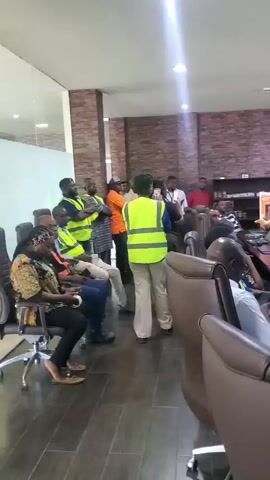 This video shows a group of people in a meeting room. Most of the people are wearing high-visibility vests, suggesting they might be at a work-related event or in a safety-conscious environment. There is a large conference table in the center of the room, with laptops and bottles of water on it. Some individuals are seated at the table, while others are standing. The room appears to be an office or conference space, with brick-patterned walls and blinds on the windows. The lighting is artificial, coming from overhead fixtures. The overall atmosphere appears to be one of instruction or discussion, as people are looking towards the front of the room where some individuals are standing and facing the group.