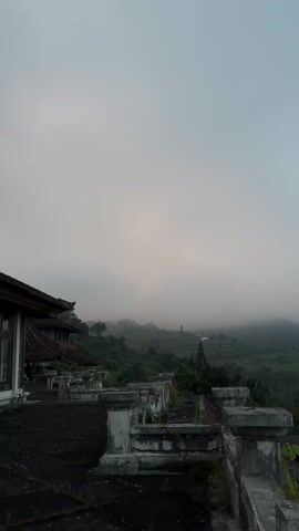 The video shows a misty mountain landscape viewed from a dilapidated building's balcony. The building has a tile roof and weathered, overgrown concrete railings. In the distance, rolling hills are covered with dense green vegetation and shrouded in thick fog. The sky is overcast, contributing to the subdued, ethereal atmosphere.