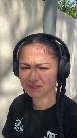 A medium shot depicts a person with dark hair styled in two braids, wearing black headphones and a black t-shirt, standing outdoors against a light-colored textured wall. They are squinting with a pained expression. There is a pole to the right of the person and some greenery visible in the background. The light is bright and sunny. The person is looking directly at the camera.