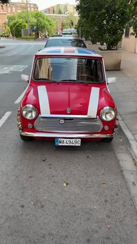 A red Mini Cooper is parked on the side of a street. The car has white stripes on the hood and a Union Jack flag on the roof. The license plate reads "MA 4949C". There is a black car parked behind it. The street is paved and there are some fallen leaves on the ground. There are trees and buildings visible in the background.