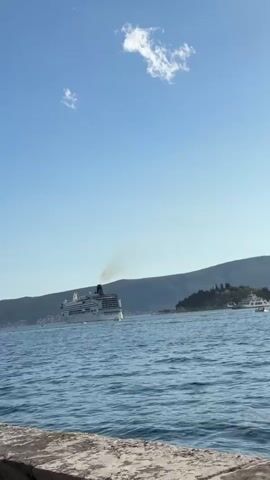 This media file captures a serene daytime scene on a body of water, likely a bay or inlet, in Lepetane, Montenegro. The primary subject is a large, white cruise ship sailing across the calm blue water. The ship is emitting a plume of dark smoke from its funnel, indicating it is in operation. In the background, a lush, green hillside rises from the water's edge, dotted with trees and some buildings. The sky is a clear, bright blue with a few wispy white clouds.

To the left of the cruise ship, several smaller boats are visible, some appearing to be moored or anchored. In the foreground, a stone or concrete embankment lines the edge of the water, suggesting a quay or a waterfront promenade. The lighting suggests it is a sunny day, likely late morning or early afternoon, given the brightness and the shadows. The overall atmosphere is peaceful and idyllic, conveying a sense of travel and scenic beauty. There is no visible text in the media file.