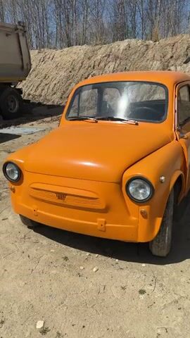 An orange vintage car is parked outside on a sunny day. The car is in a dirt parking lot. Behind the car is a large pile of dirt. Behind the dirt pile is a forest of trees. To the right is a large yellow trailer.