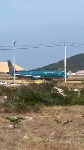 The video shows a Vietnam Airlines airplane on a runway. The airplane is light blue and has the words "Vietnam Airlines" written in white. The plane is moving at a moderate pace. The background includes a clear blue sky, green hills, power lines, and light poles.  There is scrubby, dry vegetation in the foreground. The wind is moderately strong.