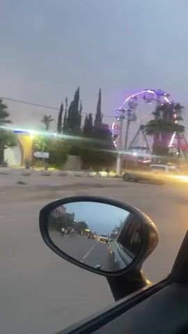 The video shows a drive past an amusement park during dusk or early evening, seen from the passenger side window of a moving car. The sky is overcast, and artificial lights are visible at the amusement park.

In the initial frames, a large white van with blurred blue text on its side passes by on the left. The car's side mirror prominently reflects the road behind, showing other vehicles and distant buildings.

As the car moves forward, a vibrant amusement park comes into view on the right. Key attractions visible include a large Ferris wheel with colorful gondolas, and a prominent pirate ship ride with the text "PIRATES LIFE" illuminated on its structure. Several palm trees are also part of the scenery. Further down, a brightly lit carousel or merry-go-round, blurred due to motion and its numerous lights, is seen.

The area around the amusement park has many parked cars, suggesting activity and visitors. People are also seen walking on the sidewalks. The overall impression is of a lively evening scene at an entertainment venue.