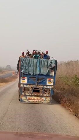 From a moving vehicle, the video shows the rear of a blue truck driving on a paved road. Multiple people are riding on top of the truck, some waving. The truck has a blue tarp covering the top of its load. The road is bordered by dry, grassy vegetation. Another vehicle is visible in the distance. The sky is overcast.