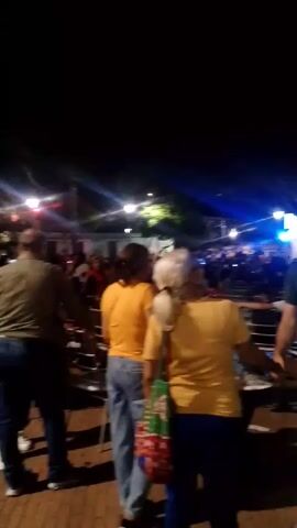 The video depicts an outdoor nighttime gathering. A crowd is present, illuminated by stage lights. The camera focuses on a group of individuals; one man embraces another. The environment consists of trees and open space, suggesting a park. The scene includes people of various ages and ethnicities, some walking and others standing still.