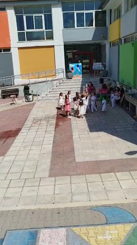 A group of young children in white dresses are standing on a paved courtyard outside a school building. Some of the children are holding up long, colorful cylindrical objects. A woman in a pink shirt is standing behind them, and another person is sitting on a bench to the right. The school building has a colorful facade with large windows and a set of stairs leading up to the entrance. The courtyard is paved with square tiles, and there are some colorful chalk drawings on the ground. The lighting suggests it is daytime and sunny.