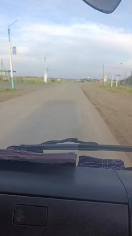 The video provides a first-person perspective from inside a vehicle, looking out through the windshield. The vehicle is traveling on a paved road that appears to be in a rural or semi-rural area. The road is mostly empty, with a few signs and structures visible in the distance. The sky is cloudy, suggesting either early morning or late afternoon. The dashboard of the vehicle is partially visible in the foreground, indicating the camera is mounted inside. There are no people or other vehicles clearly visible on the road or in the immediate surroundings. The overall impression is of a calm and uneventful journey.