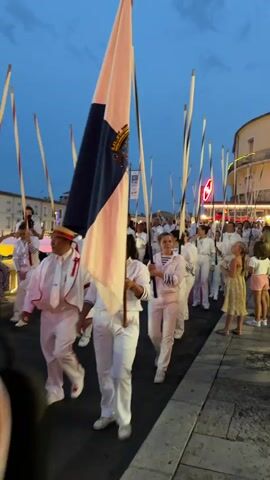 A group of people in white outfits with pink trim are marching in what appears to be a parade or ceremonial event. They are carrying tall, thin poles with white and black banding near the top. Many of the people are holding the poles upright, and some are carrying a large flag with blue and pink sections and a crest. They are walking on a paved surface, likely a street or sidewalk, with buildings and streetlights visible in the background. The sky is a deep blue, indicating it is dusk or early evening. Some spectators are visible on the right side of the frame.