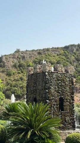 A stone tower stands in the foreground with water spraying from its top. In front of the tower, a palm tree is visible. Behind the tower, a hillside is covered with green vegetation. Multiple fountains spray water to the right of the tower. The sky is clear and blue.