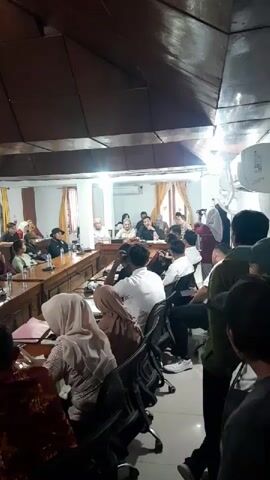 This is a video of a meeting in a room with many people. The room has a long wooden table with chairs around it. There are papers and microphones on the table. Some people are sitting at the table and some are standing. The people at the table are talking and listening to each other. The people standing are observing the meeting. The room has wooden walls and ceiling and is lit by artificial lights. There are windows in the room with curtains. The video is shaky and not very clear.