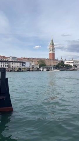 The camera is pointed at the shore of Venice, Italy, from a boat in the water. The shore is lined with buildings, including the St. Mark's Campanile, and docked boats. The water is choppy with small waves and a few boats are moving in the distance. The sky is partly cloudy. Part of the boat the camera is on is visible in the bottom left corner of the frame. A child is heard speaking in the background.